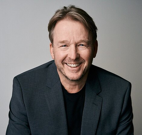 Smiling man in dark blazer and black shirt against neutral background