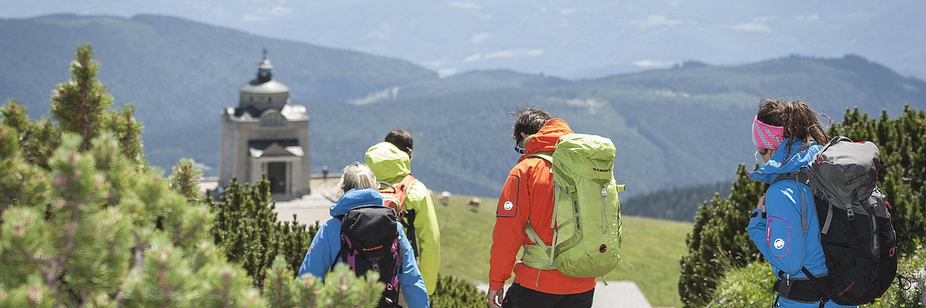 Gruppe von Wanderern mit Rucksäcken auf Bergpfad in Richtung einer Alpenkapelle